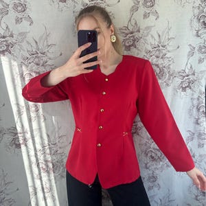 Puede incluir: Una chaqueta roja con botones dorados y costuras decorativas. La chaqueta tiene mangas largas y una silueta ajustada.