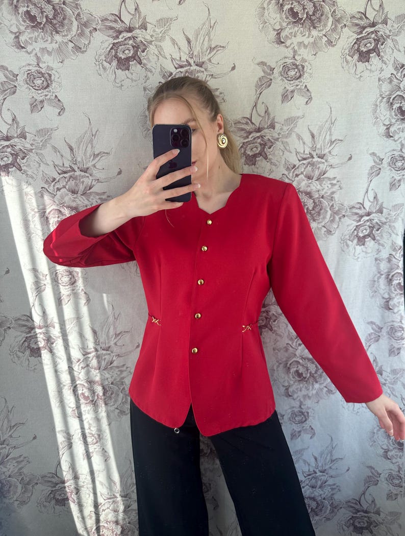 Puede incluir: Una chaqueta roja con botones dorados y costuras decorativas. La chaqueta tiene mangas largas y una silueta ajustada.