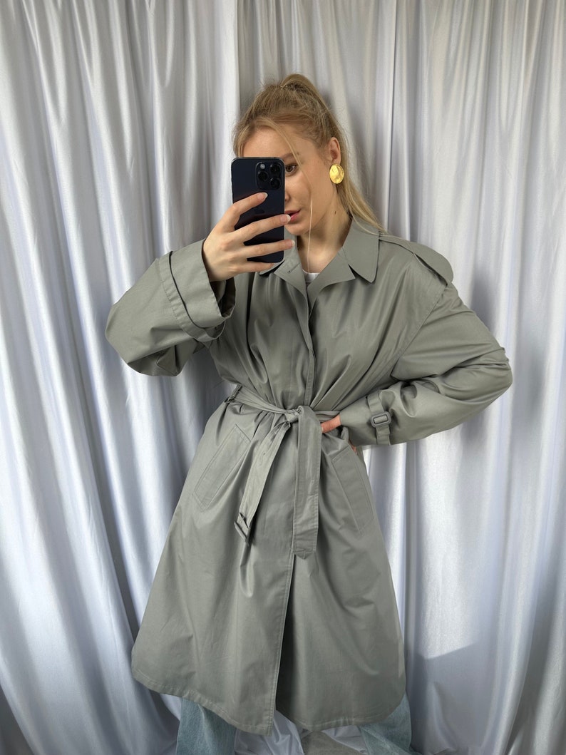 May include: A light gray trench coat with a belt and a button closure. The coat has a classic design and is made of a lightweight fabric.