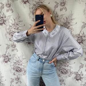 Pode incluir: Uma mulher vestindo uma camisa branca e azul listrada com bot&otilde;es e gola de renda e jeans azul claro.