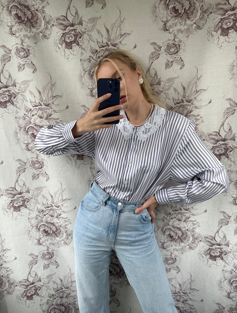 Pode incluir: Uma mulher vestindo uma camisa branca e azul listrada com bot&otilde;es e gola de renda e jeans azul claro.
