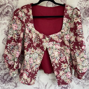May include: A maroon floral print cropped jacket with a square neckline and puffed sleeves. The jacket has a single button closure at the front.