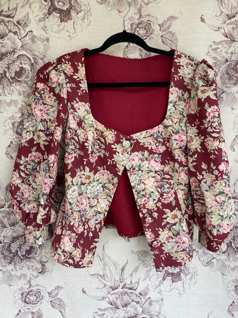 May include: A maroon floral print cropped jacket with a square neckline and puffed sleeves. The jacket has a single button closure at the front.