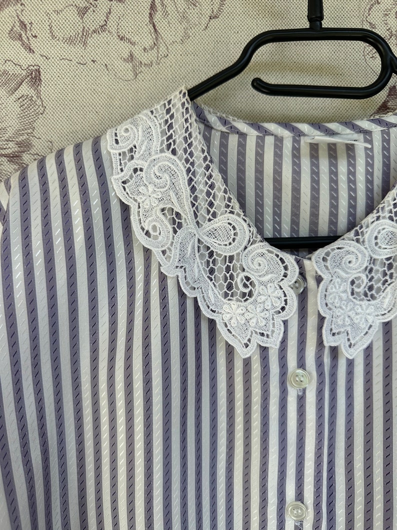 Pode incluir: Uma blusa listrada branca e roxa com uma gola de renda branca. A gola tem uma borda festonada e um design floral.