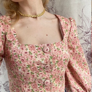 May include: A pink floral cropped top with a square neckline and five button closures. The top has a delicate floral pattern with pink, white, and green flowers.
