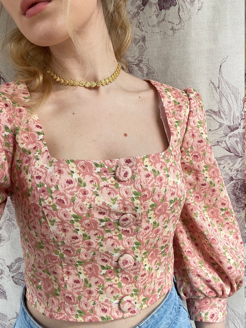 May include: A pink floral cropped top with a square neckline and five button closures. The top has a delicate floral pattern with pink, white, and green flowers.