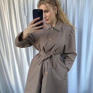 May include: A brown belted trench coat with a button closure and a collar. The coat has a cinched waist and long sleeves.