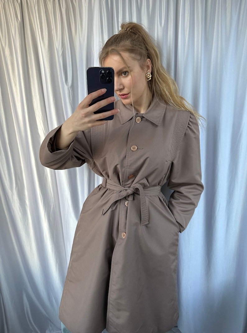 May include: A brown belted trench coat with a button closure and a collar. The coat has a cinched waist and long sleeves.