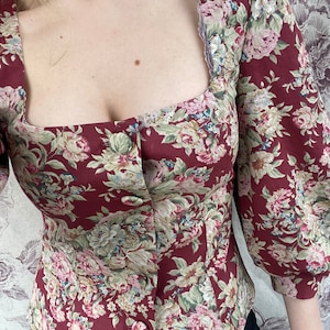 May include: A maroon floral print blouse with a square neckline and button closure. The blouse has 3/4 sleeves and a fitted silhouette.