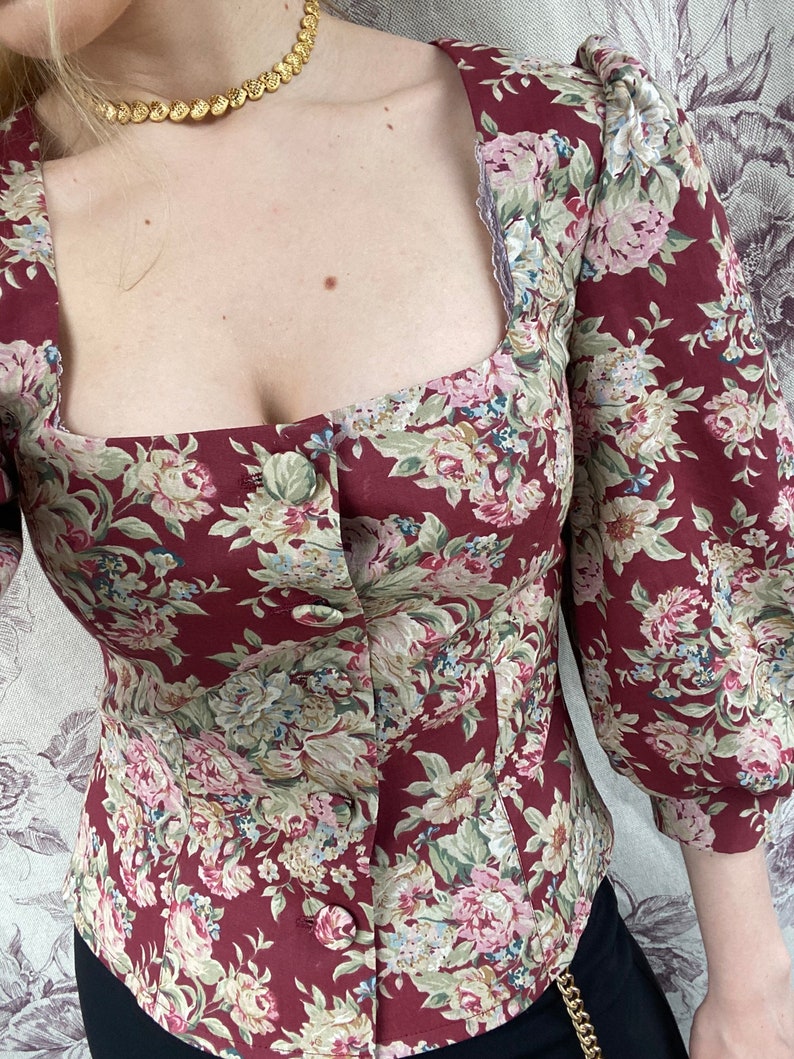 May include: A maroon floral print blouse with a square neckline and button closure. The blouse has 3/4 sleeves and a fitted silhouette.