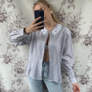 Pode incluir: Uma camisa de manga comprida listrada branca e roxa com uma gola de renda branca. A camisa est&aacute; desabotoada e tem um caimento solto.
