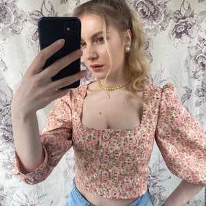 May include: A pink floral print cropped top with a square neckline and puff sleeves. The top has a button closure in the front and is made of a lightweight fabric.