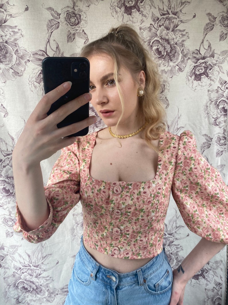 May include: A pink floral print cropped top with a square neckline and puff sleeves. The top has a button closure in the front and is made of a lightweight fabric.