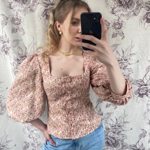 May include: A pink floral print blouse with puffed sleeves and a square neckline. The blouse has a button closure and is fitted at the waist.