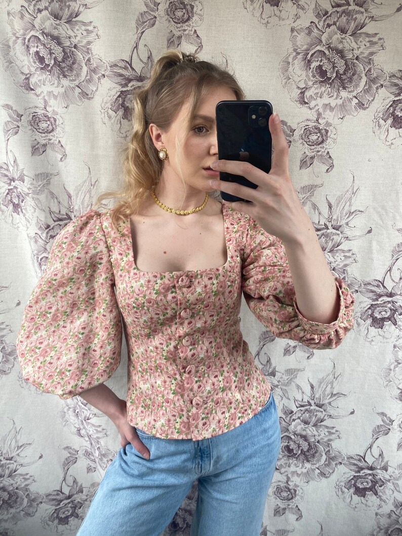 May include: A pink floral print blouse with puffed sleeves and a square neckline. The blouse has a button closure and is fitted at the waist.