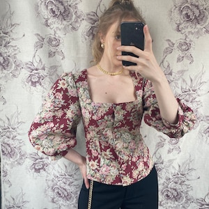 May include: A red and white floral print blouse with a square neckline and puffed sleeves. The blouse has a button closure at the front and is cinched at the waist. The model is wearing black pants with a gold chain belt.