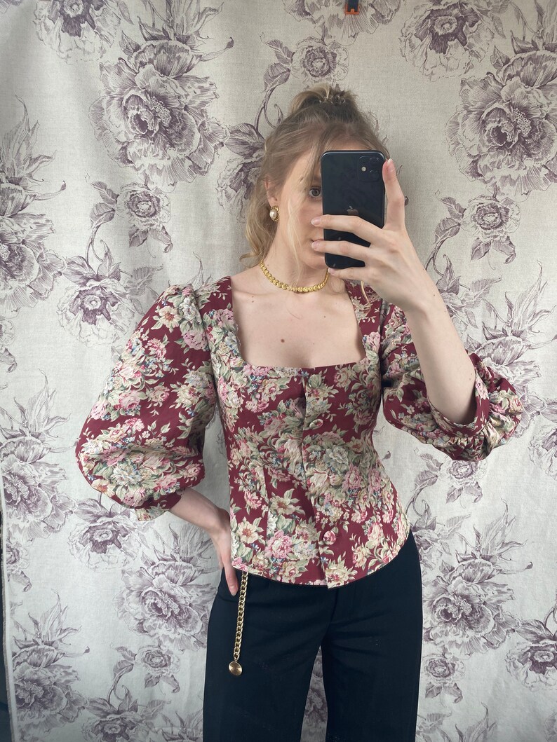 May include: A red and white floral print blouse with a square neckline and puffed sleeves. The blouse has a button closure at the front and is cinched at the waist. The model is wearing black pants with a gold chain belt.