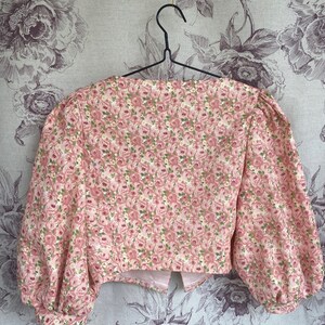May include: A pink cropped blouse with a floral print featuring small pink roses and green leaves. The blouse has a square neckline and puffed sleeves.