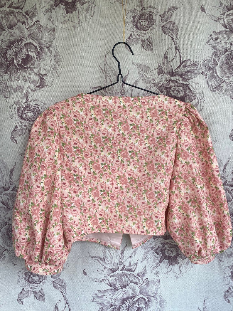 May include: A pink cropped blouse with a floral print featuring small pink roses and green leaves. The blouse has a square neckline and puffed sleeves.