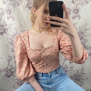 May include: A pink floral print cropped blouse with puffed sleeves and a square neckline. The blouse has a button closure in the front and is paired with light blue denim jeans.