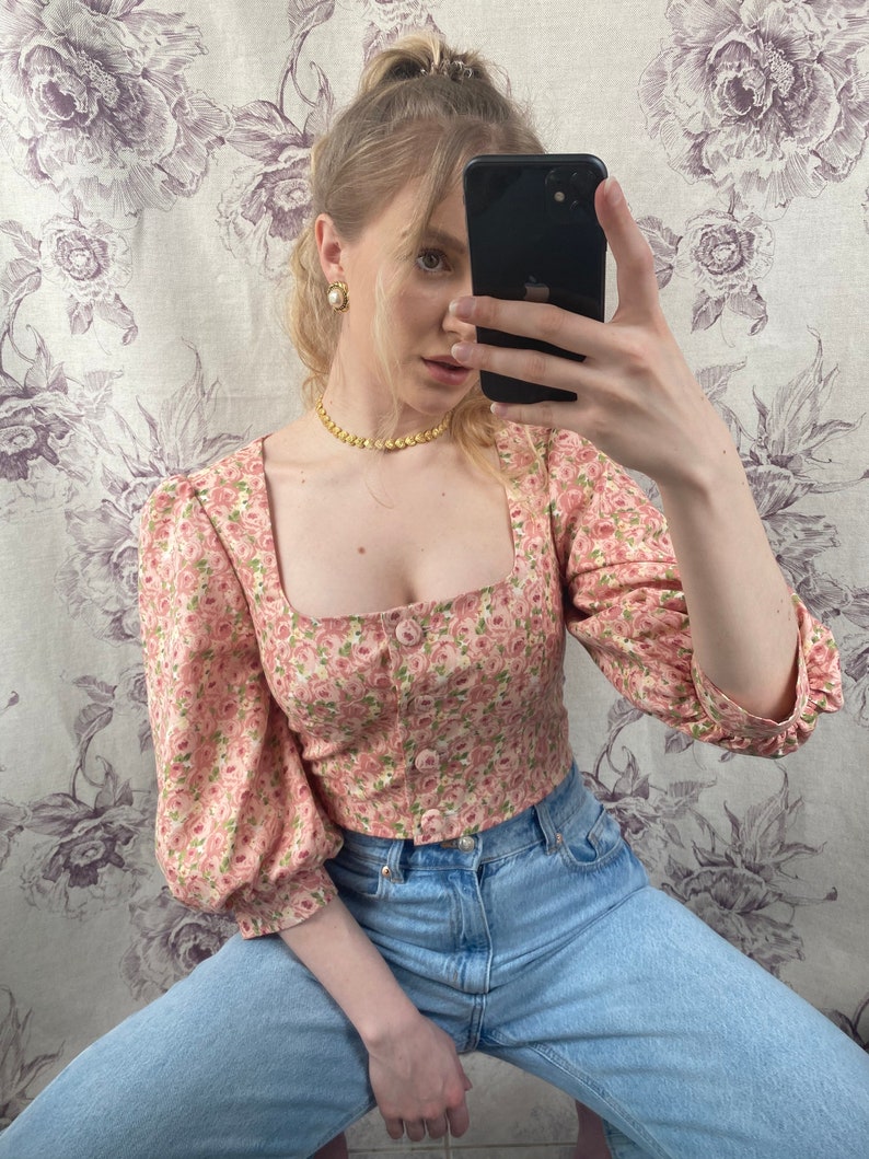 May include: A pink floral print cropped blouse with puffed sleeves and a square neckline. The blouse has a button closure in the front and is paired with light blue denim jeans.