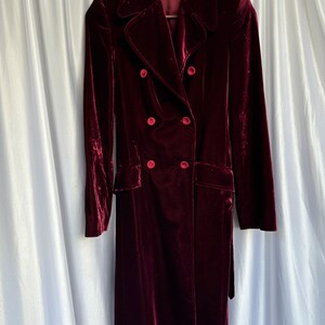 May include: A long, burgundy velvet coat with a double-breasted closure and a belt. The coat has four large buttons and a classic collar.