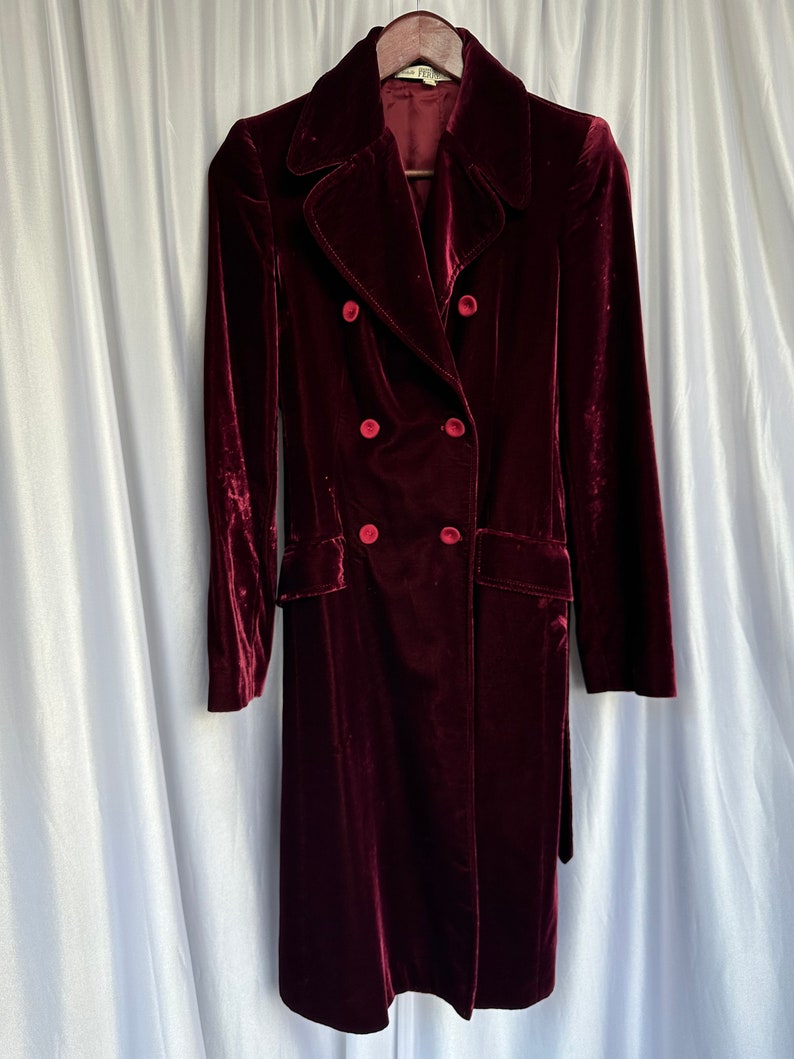 May include: A long, burgundy velvet coat with a double-breasted closure and a belt. The coat has four large buttons and a classic collar.