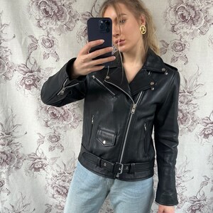 May include: A black leather motorcycle jacket with a zipper closure and a belt at the waist. The jacket has a  stand-up collar and long sleeves.