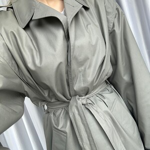 May include: A light gray trench coat with a belt and a button closure. The coat has a classic design and is made of a lightweight fabric.