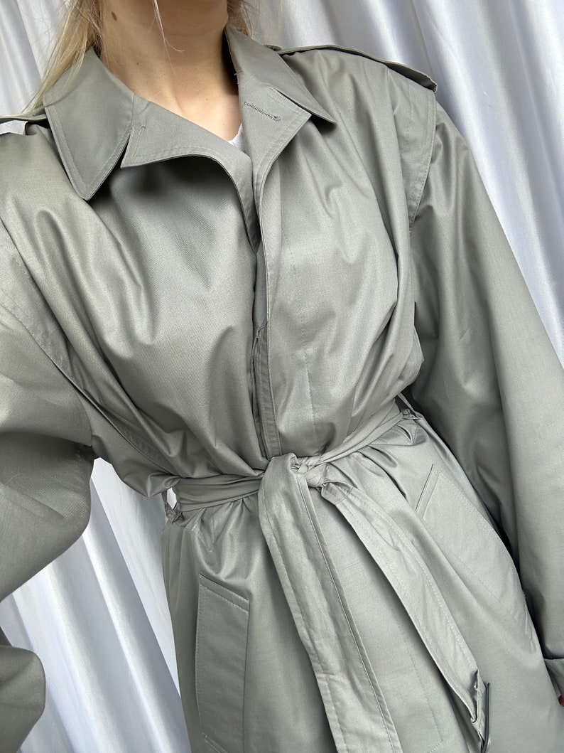 May include: A light gray trench coat with a belt and a button closure. The coat has a classic design and is made of a lightweight fabric.