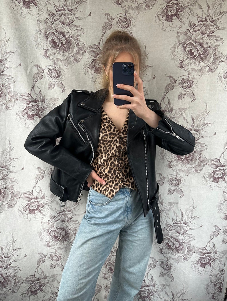 May include: A woman wearing a black leather jacket with a brown and black leopard print shirt and light blue jeans.