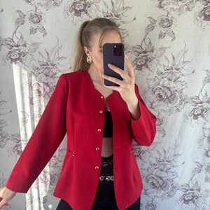 Puede incluir: Una chaqueta roja con botones dorados y un detalle de cadena en las costuras laterales. La chaqueta es corta y tiene un corte relajado.