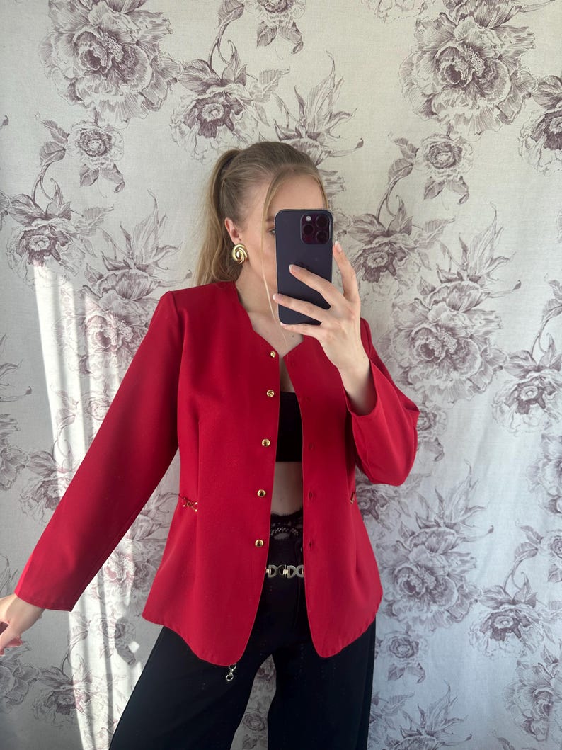 Puede incluir: Una chaqueta roja con botones dorados y un detalle de cadena en las costuras laterales. La chaqueta es corta y tiene un corte relajado.