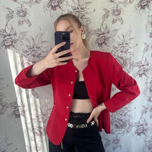 Puede incluir: Una chaqueta roja con botones dorados y un cintur&oacute;n de cadena dorada. La chaqueta tiene mangas largas y cuello en V.