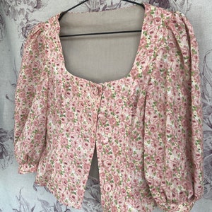 May include: A pink and white floral print blouse with a square neckline and puffed sleeves. The blouse has a button closure in the front.