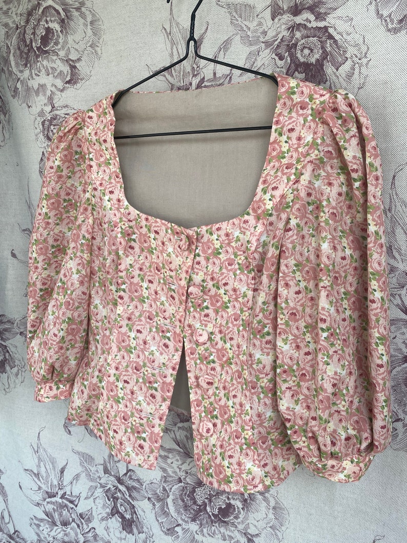May include: A pink and white floral print blouse with a square neckline and puffed sleeves. The blouse has a button closure in the front.