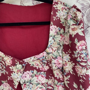 May include: A maroon floral print jacket with a square neckline and a single button closure. The jacket has a fitted bodice and puffed sleeves.