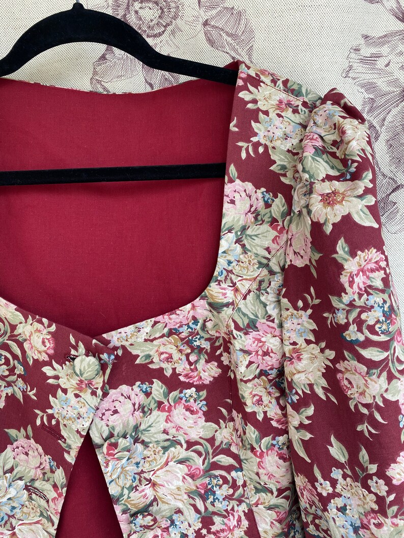 May include: A maroon floral print jacket with a square neckline and a single button closure. The jacket has a fitted bodice and puffed sleeves.