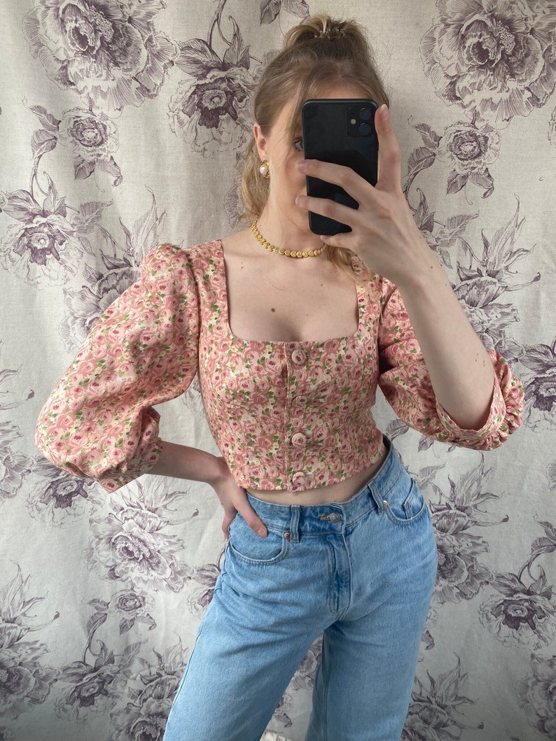 May include: A pink floral crop top with puffed sleeves and a square neckline. The top has a button closure in the front and is made of a lightweight fabric.
