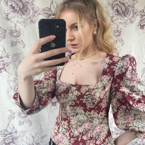 May include: A woman wearing a fitted, maroon floral print top with puffed sleeves and a square neckline. She is wearing a gold chain necklace and gold earrings.