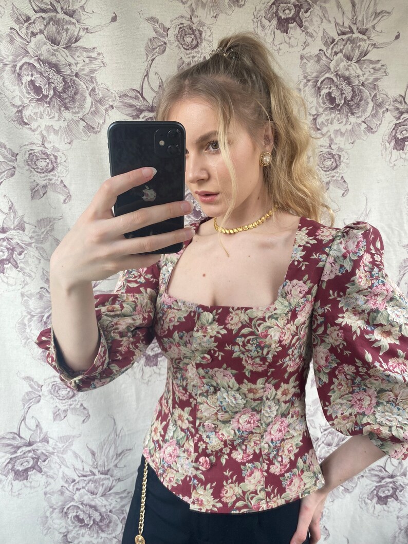 May include: A woman wearing a fitted, maroon floral print top with puffed sleeves and a square neckline. She is wearing a gold chain necklace and gold earrings.
