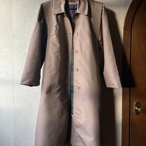 May include: A long, light brown coat with a button-up front and a collar. The coat has a plaid lining and is hanging on a hanger.