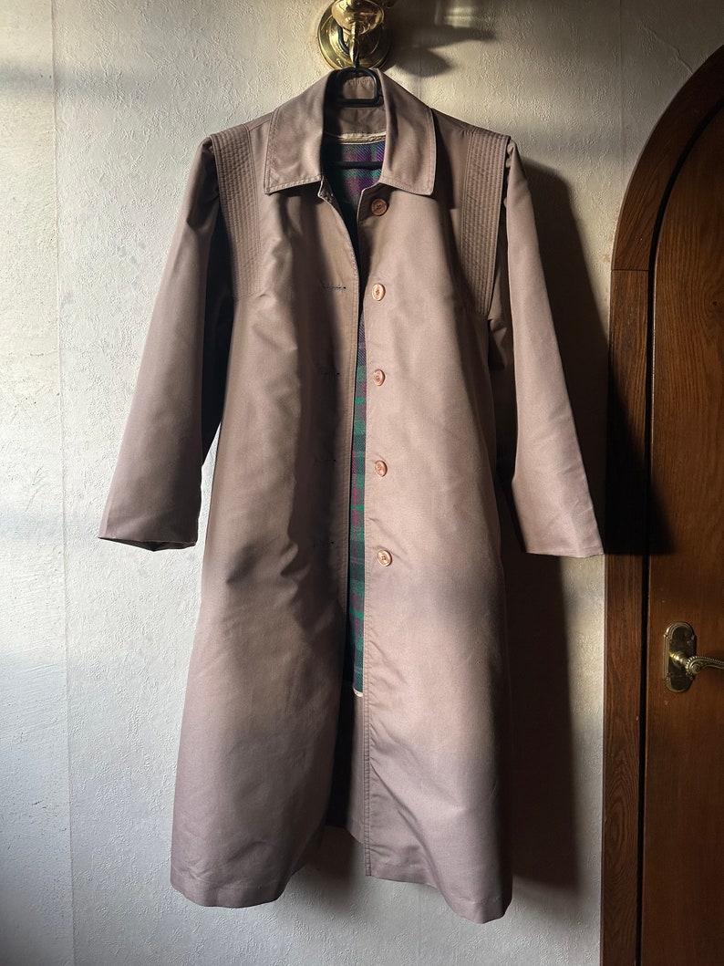 May include: A long, light brown coat with a button-up front and a collar. The coat has a plaid lining and is hanging on a hanger.