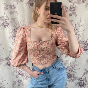 May include: A pink floral cropped top with puffed sleeves and a square neckline. The top has a button closure and is paired with light blue jeans.