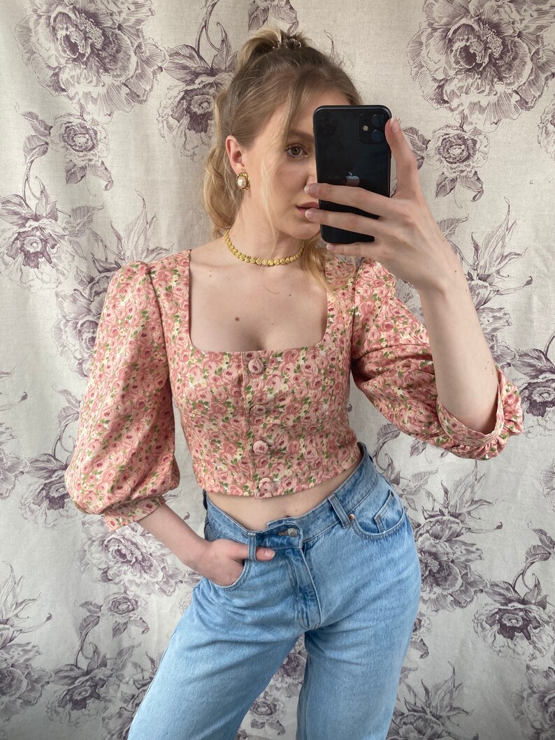 May include: A pink floral cropped top with puffed sleeves and a square neckline. The top has a button closure and is paired with light blue jeans.