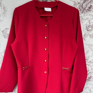 Puede incluir: Una chaqueta roja con botones dorados y costuras decorativas en los lados. La chaqueta tiene cuello en V y mangas largas.