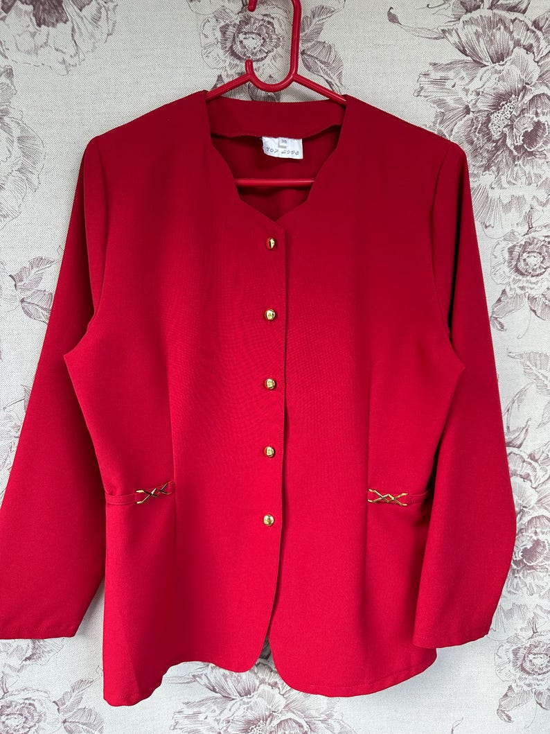 Puede incluir: Una chaqueta roja con botones dorados y costuras decorativas en los lados. La chaqueta tiene cuello en V y mangas largas.