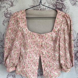 May include: A pink floral print blouse with a square neckline and a single button closure. The blouse has puffed sleeves and a cropped length.