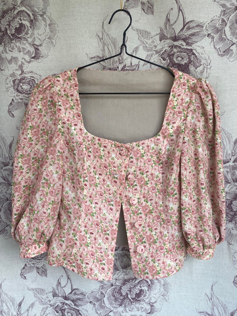 May include: A pink floral print blouse with a square neckline and a single button closure. The blouse has puffed sleeves and a cropped length.