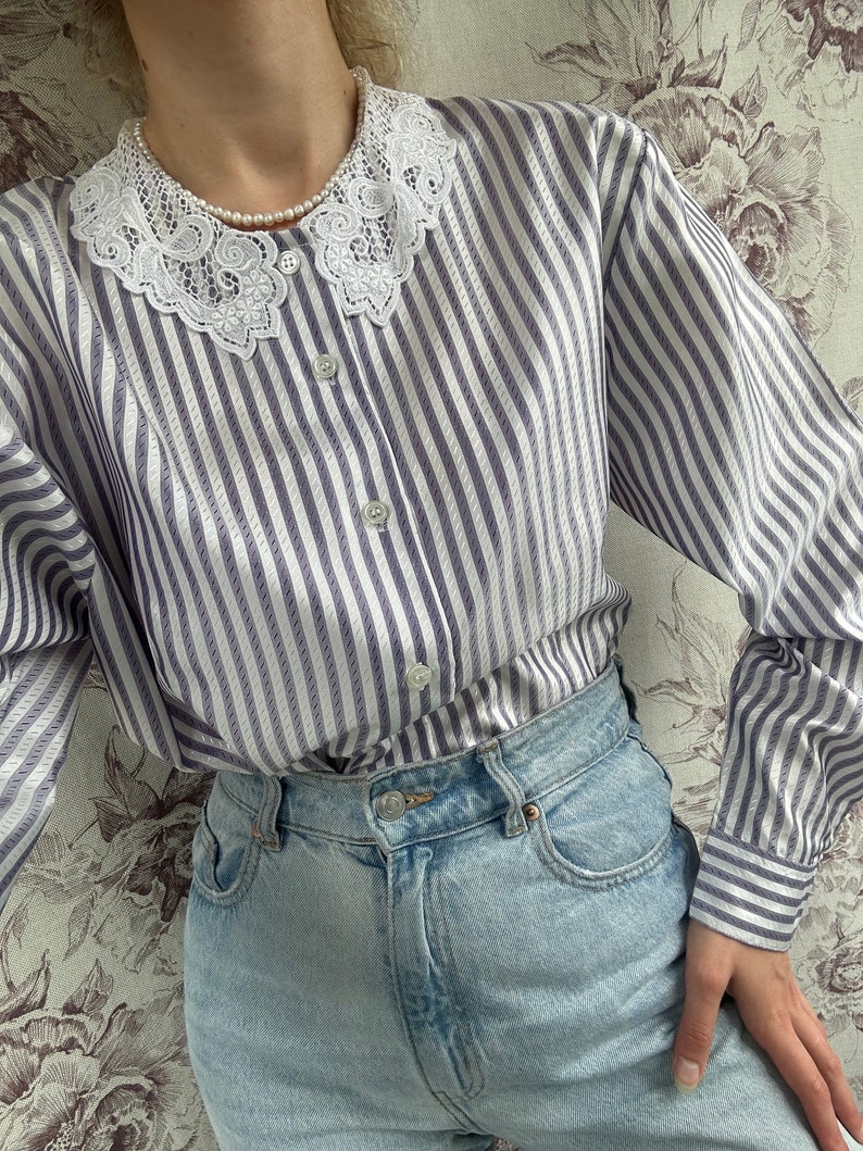 Pode incluir: Uma camisa de manga comprida listrada com uma gola de renda branca. A camisa &eacute; de cor roxo claro e branco. A camisa est&aacute; enfiada em uma cal&ccedil;a jeans azul claro.